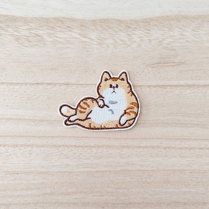May include: Embroidered iron-on patch featuring a cartoon orange tabby cat lying on its side with its paw on its belly.