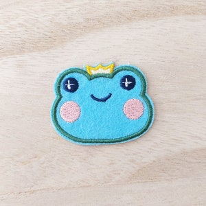 May include: A blue felt frog with a yellow crown, embroidered with pink cheeks and sparkling eyes.