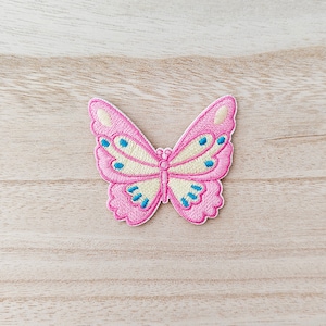 May include: A pink butterfly embroidered patch with yellow wings and blue accents. The patch is on a light brown wooden surface.