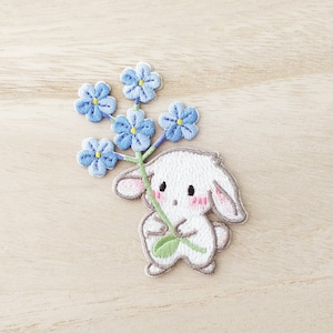 May include: Embroidered patch of a white bunny holding blue forget-me-nots. The bunny has pink inner ears and cheeks, with a grey outline. The flowers have yellow centres. The patch is on a light wood grain background.