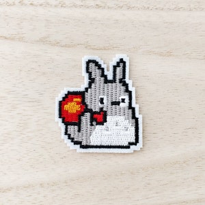 May include: Embroidered patch featuring a pixelated gray bunny holding a red object. The bunny has black outlines and white details. The patch is on a light wood grain surface.