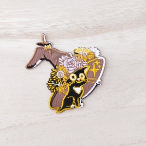 May include: A black cat wearing a brown witch hat with yellow and white flowers. The hat has a brown brim and a white band around the top. The cat has white markings on its chest and a white nose.