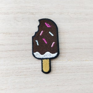 May include: A brown chocolate popsicle with a bite taken out of it. The popsicle is covered in white and pink sprinkles and has a yellow stick.