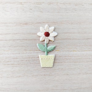 May include: A white flower with a red centre is embroidered on a green stem and leaves. The flower is in a yellow pot.