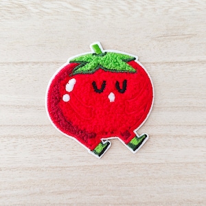 May include: Embroidered patch featuring a smiling red tomato with green leaves and black eyes. The tomato has small legs with green and black details. The patch has a white border and is designed for application on fabric.