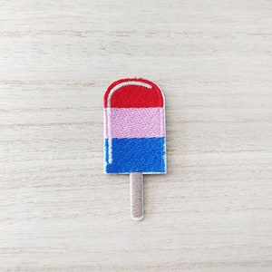 Peut inclure: Un patch brodé en forme de popsicle rouge, rose et bleu avec un bâton blanc.