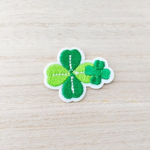 May include: A green four-leaf clover embroidered patch with white stitching. The patch is on a light wood background.