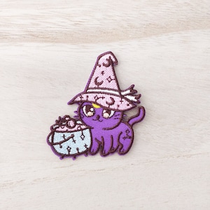 May include: Embroidered patch of a purple cat wearing a pink witch's hat decorated with moons and stars. The cat is beside a light blue cauldron with a bubbling potion. The patch is a whimsical design.