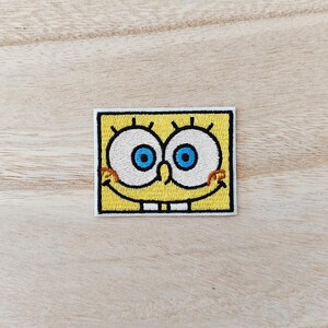May include: A rectangular embroidered patch featuring a yellow background with a character face design. The design includes two large white circular eyes with bright blue pupils, a small nose, a wide smiling mouth, and two white teeth.