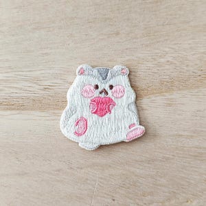 May include: Embroidered patch with a cartoon hamster design. The hamster is mainly white with grey stripes, pink cheeks, and pink accents on its paws and a ball it is holding. The patch is on a light brown wooden surface.