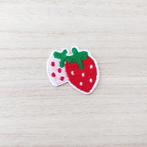 May include: Two embroidered strawberry patches, one red and one pink, with green leaves. The patches are on a light wood background.