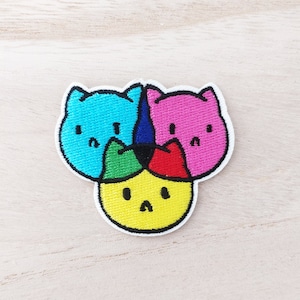 May include: An embroidered patch featuring three cartoon faces. The faces are a blue cat, a pink cat, and a yellow cat with a red hat. The patch is ideal for adding a touch of whimsy to clothing, bags, or other accessories.