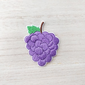 May include: Purple embroidered grape with a green stem and leaf. The grape is stitched with a quilted pattern.