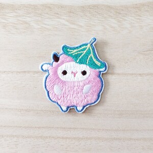 May include: Pink embroidered sheep with a green leaf on its head. The sheep has a white face with black eyes and a pink nose. The leaf is green with a white stem.