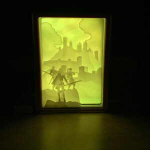May include: A light-up shadow box with a silhouette of a castle and four figures holding swords. The castle is in the background and the figures are in the foreground. The box is lit from behind, creating a glowing effect.