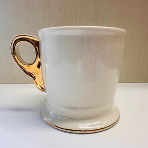 Puede incluir: Taza de afeitar de cer&aacute;mica blanca con borde y asa dorados. La taza tiene una base ancha y un cuerpo redondeado.