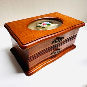 May include: A wooden jewelry box with a clear oval top, featuring a floral design. The box has two drawers with ornate metal handles. The wood has a warm, reddish-brown finish, and the box is rectangular with curved edges.
