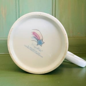 K&ouml;nnte beinhalten: Wei&szlig;e Keramiktasse mit Henkel, mit einem skurrilen Design auf der Unterseite. Das Design umfasst einen blauen Stern, einen Regenbogen und den Namen "Kinka". Der Text "&copy; 1990 RAECATH INC. LICENSEE ENESCO CORPORATION" ist ebenfalls sichtbar.