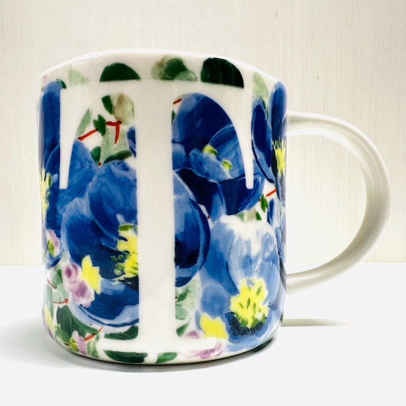 May include: White ceramic mug with a large white letter "I" on the front. The mug is decorated with blue floral patterns, green leaves, and yellow accents. The handle is white.