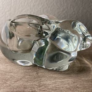 May include: A clear glass sculpture of an abstract animal, possibly a rabbit or a bear. The sculpture has a smooth, rounded surface and is designed to be used as a decorative object.