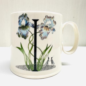 May include: A white ceramic mug featuring a floral design with blue, purple, and green irises. A black letter "I" is centered, with green leaves and two small figures at the base. The mug has a curved handle.