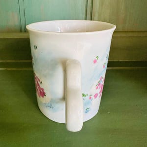 K&ouml;nnte beinhalten: Wei&szlig;e Keramiktasse mit gebogenem Henkel. Die Tasse hat ein Blumendesign mit rosa Bl&uuml;ten und blauen Akzenten. Die Signatur des K&uuml;nstlers, "Cindy", ist auf der Seite der Tasse zu sehen. Die Tasse steht vor einem gr&uuml;nen Hintergrund.