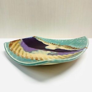 May include: A rectangular ceramic serving dish with a curved shape. The dish features a turquoise rim and a central design with purple, yellow, and brown hues. The dish is likely handmade and suitable for serving food.