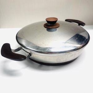 May include: A stainless steel pan with a lid and two handles. The lid has a brown wooden knob. The handles are also brown. The pan is round and sits on a white surface.