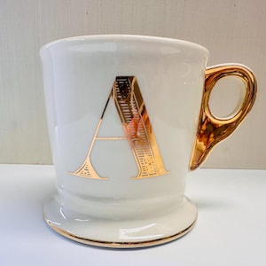 Puede incluir: Taza de cer&aacute;mica blanca con una letra A dorada en la parte delantera. La taza tiene un asa dorada.