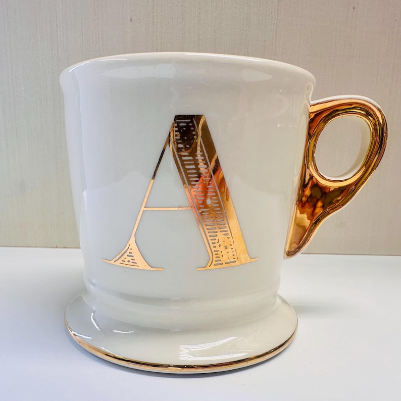 Puede incluir: Taza de cer&aacute;mica blanca con una letra A dorada en la parte delantera. La taza tiene un asa dorada.