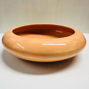 May include: A peach-colored, oval-shaped ceramic bowl with a glossy finish. The bowl has a wide, shallow design, perfect for decorative purposes or serving. The interior is a slightly darker shade of peach.