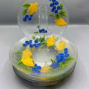 May include: A stack of eight clear glass dessert plates with a painted design of lemons and grapes. The plates have a textured surface.