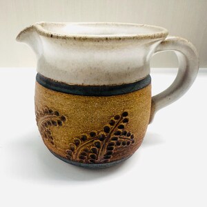 May include: A white ceramic pitcher with a brown and blue band and a leaf design. The pitcher has a handle and a spout.