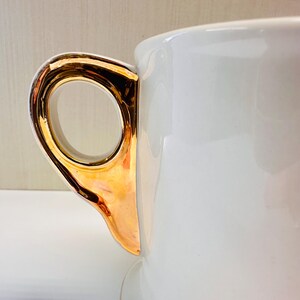 Puede incluir: Taza de t&eacute; de cer&aacute;mica blanca con un asa dorada. El asa es una simple forma de bucle.