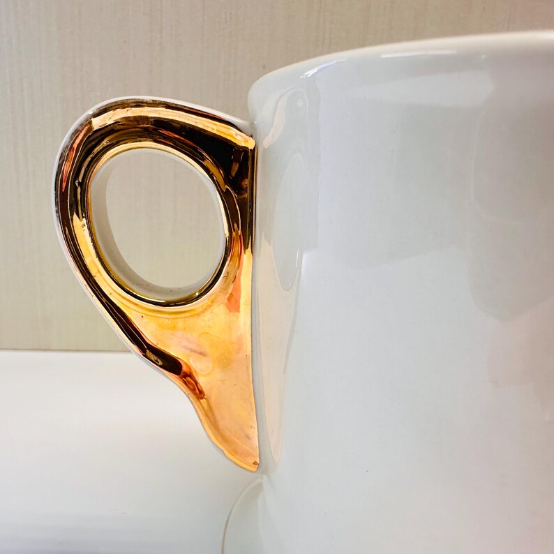 Puede incluir: Taza de t&eacute; de cer&aacute;mica blanca con un asa dorada. El asa es una simple forma de bucle.