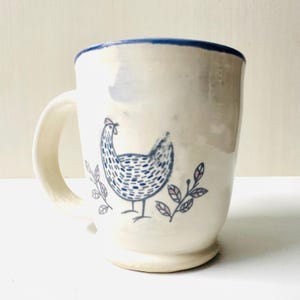 May include: A white ceramic mug with a blue rim and handle. The mug features a hand-drawn illustration of a hen in blue and white, with sprigs of leaves in pink and blue. The mug has a rounded base.