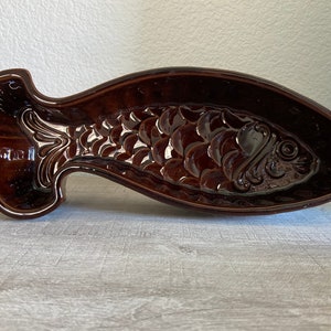 May include: A brown ceramic fish-shaped serving dish. The dish features a detailed embossed design of fish scales and facial features. The serving dish is a decorative piece for serving food.