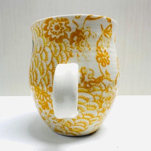 May include: A white ceramic mug with a large, curved white handle. The mug is decorated with a repeating pattern of yellow floral designs. The mug has a slightly irregular shape.