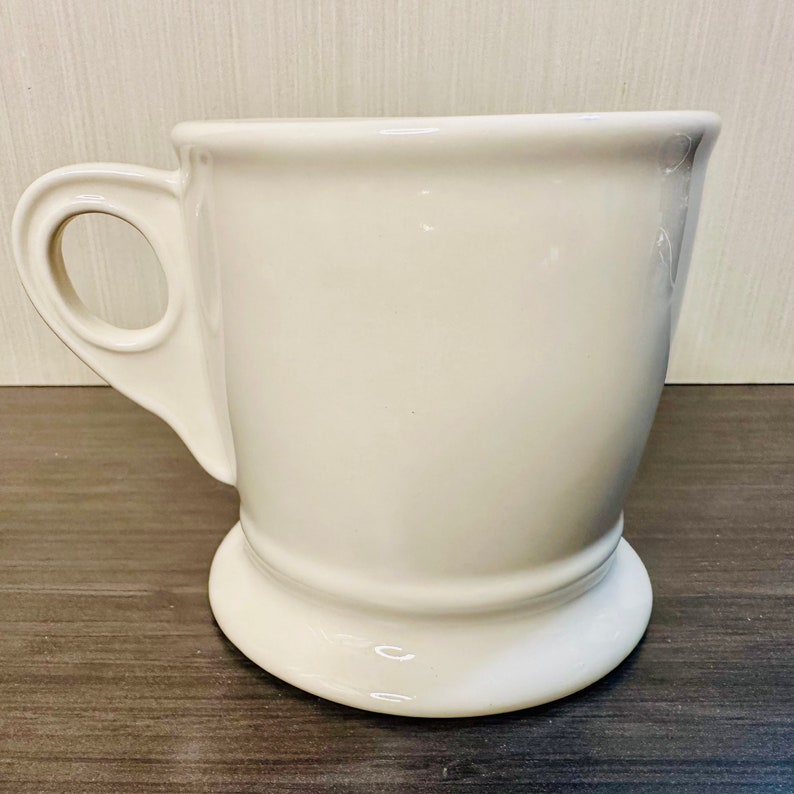 May include: A white ceramic mug with a rounded base and a handle. The mug has a simple, elegant design, suitable for tea or coffee. The handle is a classic loop shape, and the mug has a smooth, glossy finish.