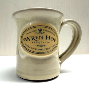 May include: A cream-colored ceramic mug with a sturdy handle and a slightly flared rim. The front features an oval-shaped tan label with dark blue text reading: SONOMA CALIFORNIA WREN HOP VINEYARDS WAKE UP & SMELL THE PINOT.