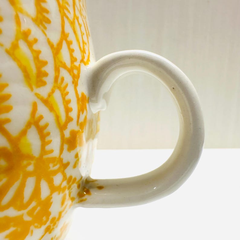 May include: A white ceramic mug with a yellow floral pattern and a curved white handle. The mug's design features a repeating pattern of stylized flowers, creating a visually appealing contrast against the white background. The handle is smooth and rounded.