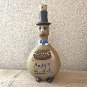 May include: A ceramic bottle shaped like a man wearing a top hat and bow tie. The bottle is brown with a brown and silver accordion in its hands. The bottle says "Andy's Scotch" on the front.