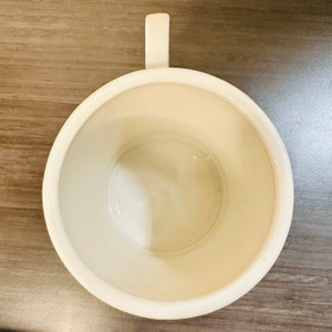May include: An overhead shot of an off-white ceramic mug with a handle. The inside of the mug is smooth and glossy. The mug is placed on a wooden surface.