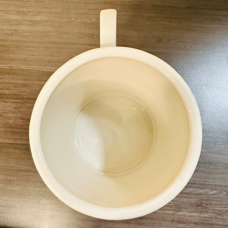 May include: An overhead shot of an off-white ceramic mug with a handle. The inside of the mug is smooth and glossy. The mug is placed on a wooden surface.