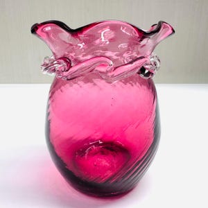 May include: A vibrant pink glass vase with a ruffled rim and a twisted glass accent. The vase has a bulbous body with a spiral design, and the glass appears translucent. The overall design is elegant and decorative.