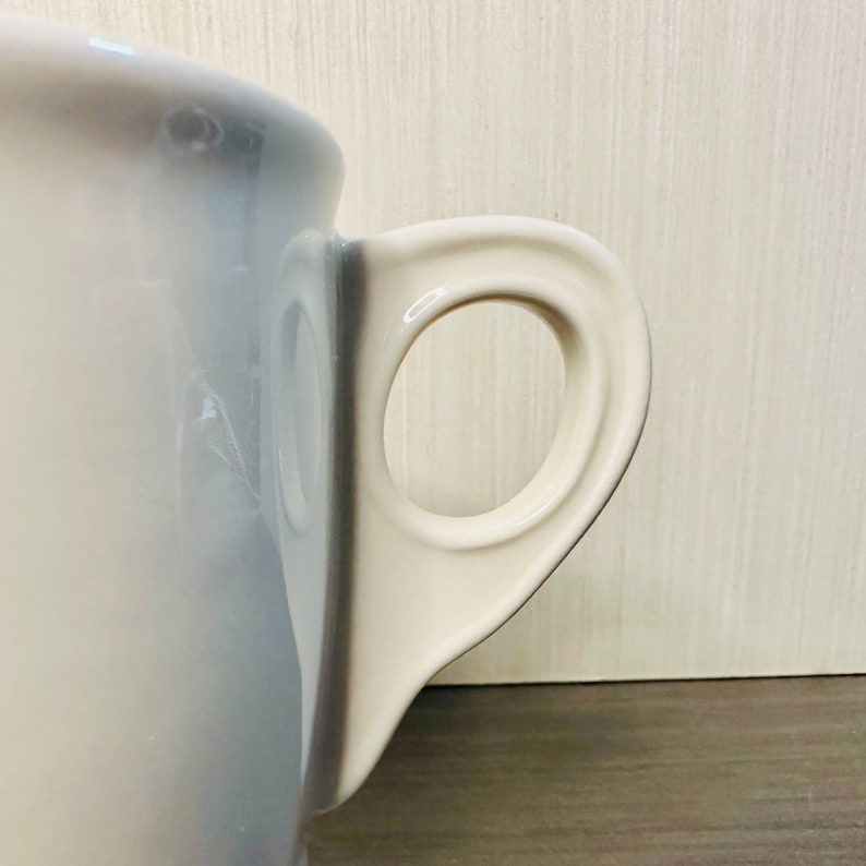 May include: Close-up of a white ceramic mug with a large, rounded handle. The mug has a glossy finish and a simple, elegant design. The handle is a contrasting off-white colour.