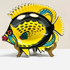 May include: A ceramic fish-shaped dish with a vibrant yellow body, black top, and yellow fins. The dish features blue and black accents, a red mouth, and a detailed eye. It is supported by a brass stand and signed "Lau Hau Ben Diller Maui 2001".