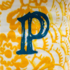 May include: Close-up of a white ceramic surface featuring a teal letter "P". The letter is centrally positioned against a backdrop of yellow floral patterns. The design has a handmade, artistic quality.