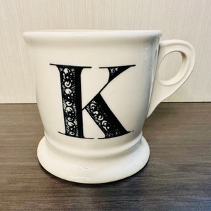 May include: White ceramic mug featuring a black letter "K". The letter is decorated with ornate patterns. The mug has a handle and a pedestal base.
