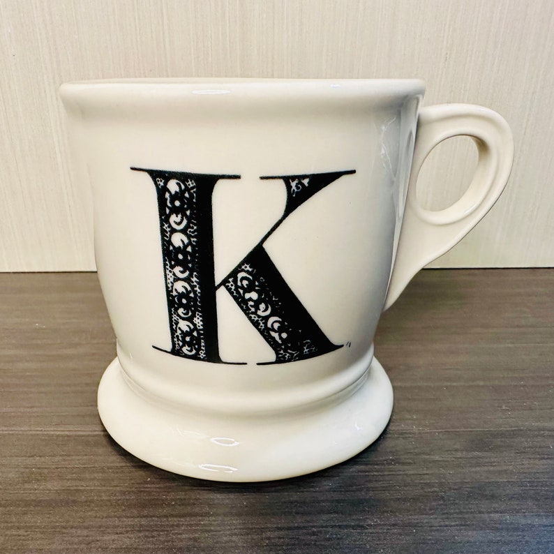 May include: White ceramic mug featuring a black letter "K". The letter is decorated with ornate patterns. The mug has a handle and a pedestal base.